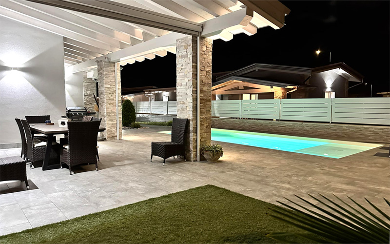 Jardin con porche de madera y barbacoa y piscina de lamina armada de obra con playa color crema y cesped artificial de noche